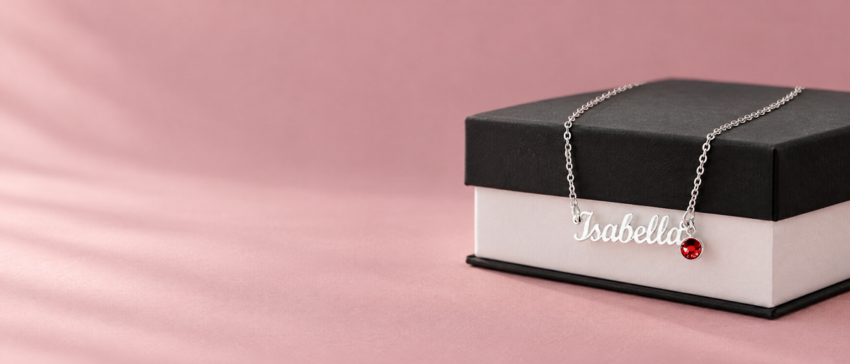 Silver name necklace with a red birthstone charm draped over a black and white gift box